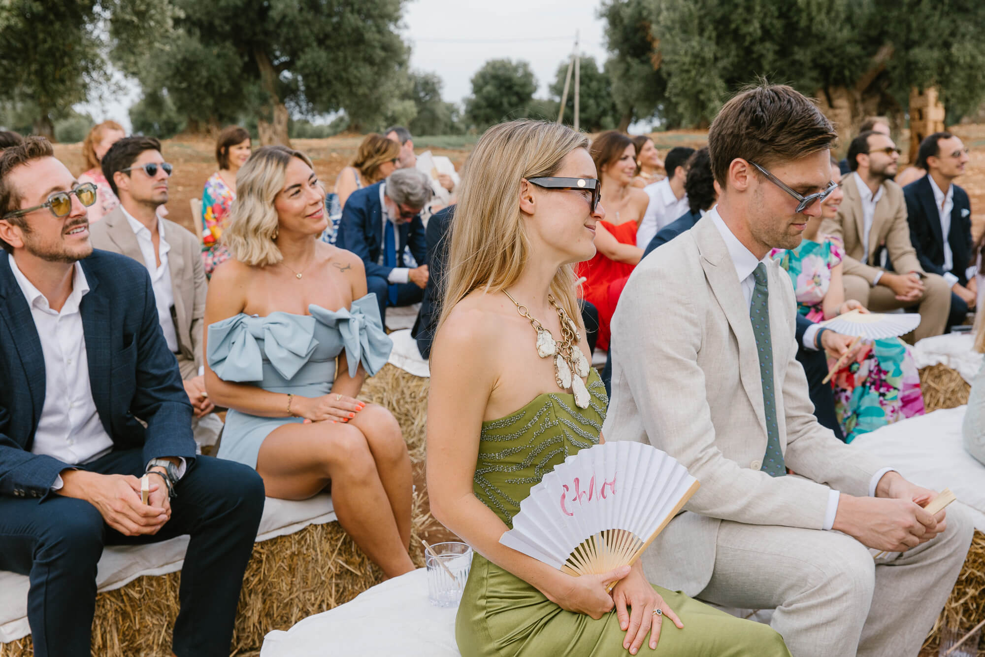 Guests on straw bales holding a wedding fan in the olive grove